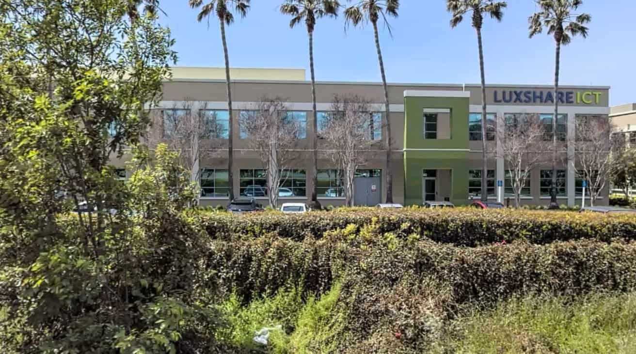 Commercial building with Luxshare ICT sign, surrounded by palm trees, greenery, and parked cars, under a clear blue sky.