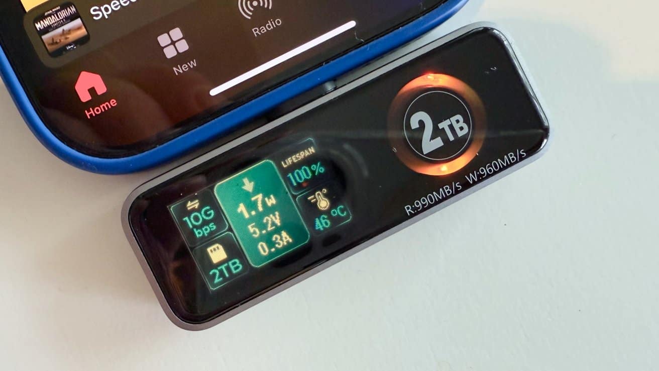 Close-up of a digital storage device with a 2TB capacity next to a smartphone. Display shows speed, power, voltage, current, lifespan, and temperature information.