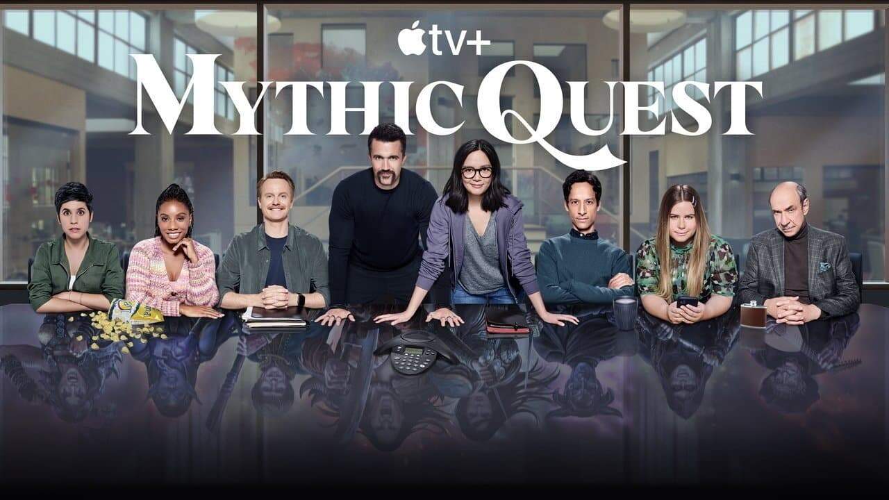 Nine people sitting at a table in a modern office setting with large white text overhead spelling 'Mythic Quest' and reflections visible on the glossy surface.
