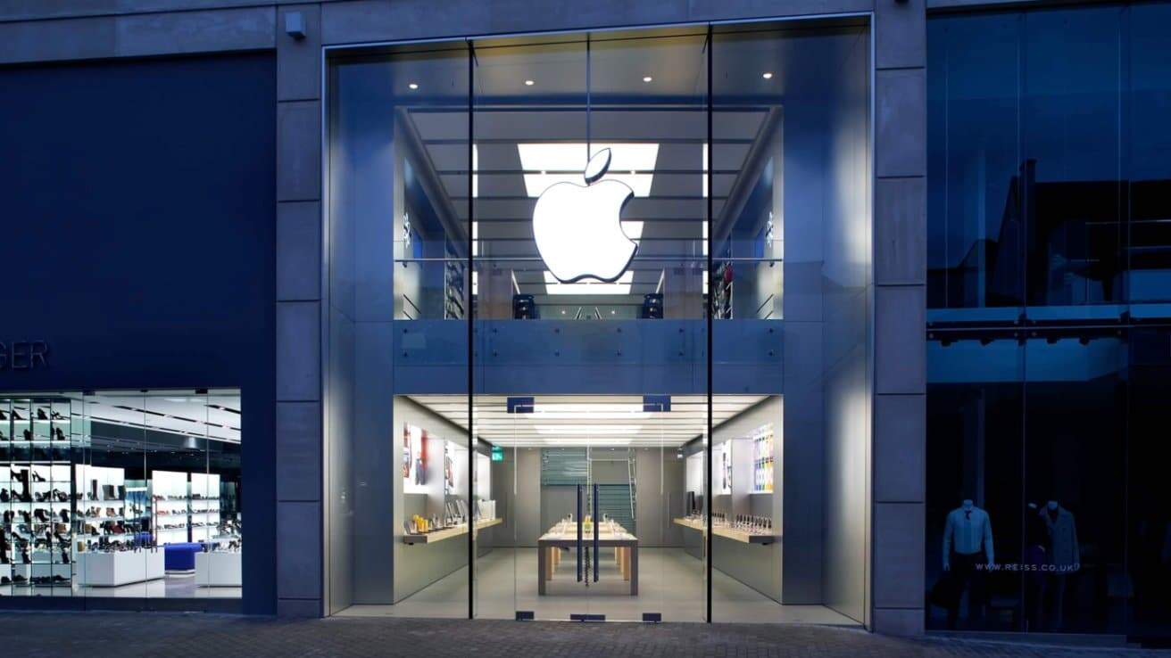 Exterior view of an Apple Store with large glass facade, prominent Apple logo, and visible interior display tables. Neighboring fashion store on the left side.