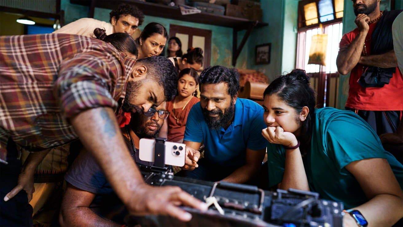 Group of people gathered around a smartphone, intently looking at its screen, in a dimly lit room filled with various objects.