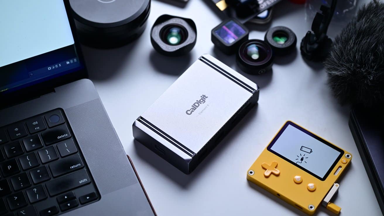A laptop keyboard, a silver device, camera lenses, and a small yellow handheld console with a low battery icon on a desk.