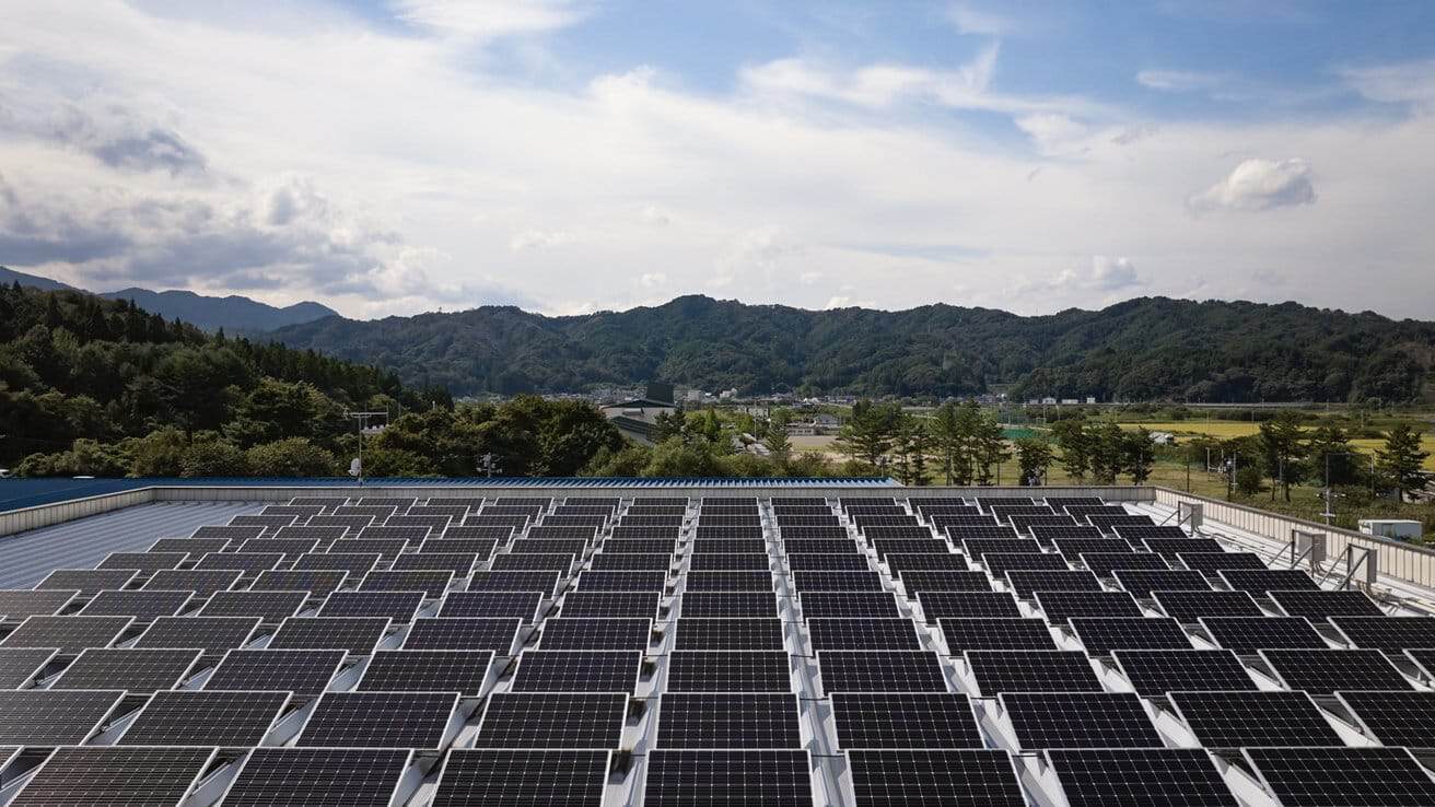 Rows of solar panels on a rooftop with a scenic mountain and forest backdrop under a partly cloudy sky.