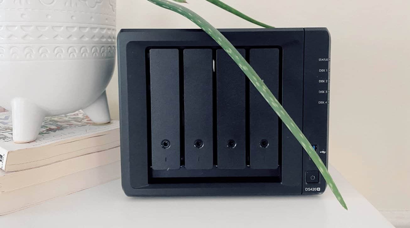 A black, four-bay network storage device next to a white decorative pot on books, with a green plant leaf extended over it.