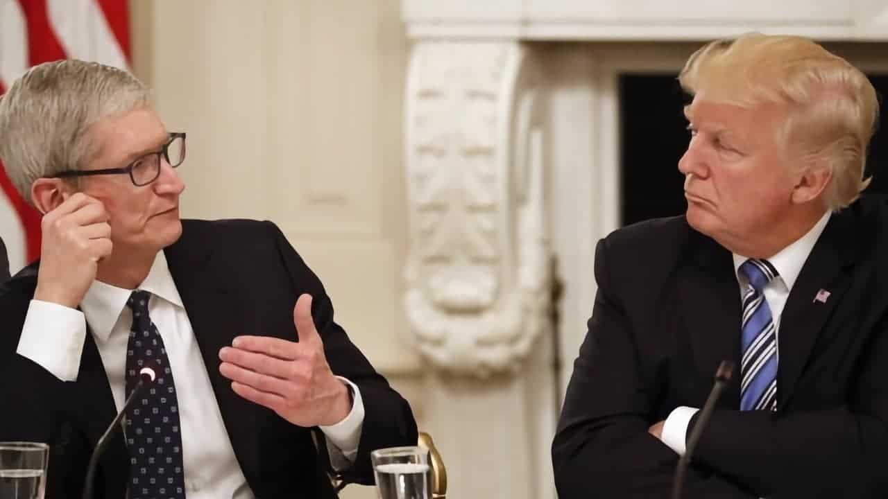 Two men in suits sit at a table, engaged in conversation against a formal setting with a white ornate background.