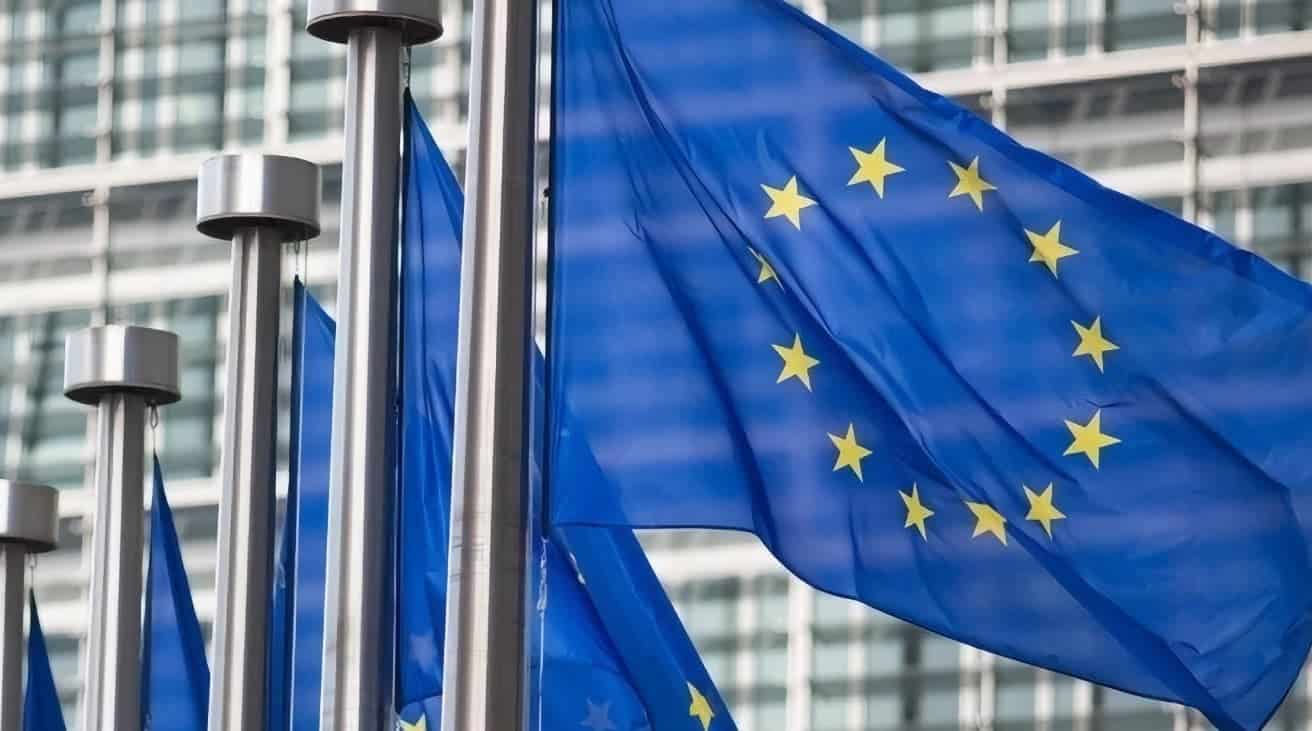 Blue flags with yellow stars wave on flagpoles, representing the European Union, set against a modern glass building backdrop.