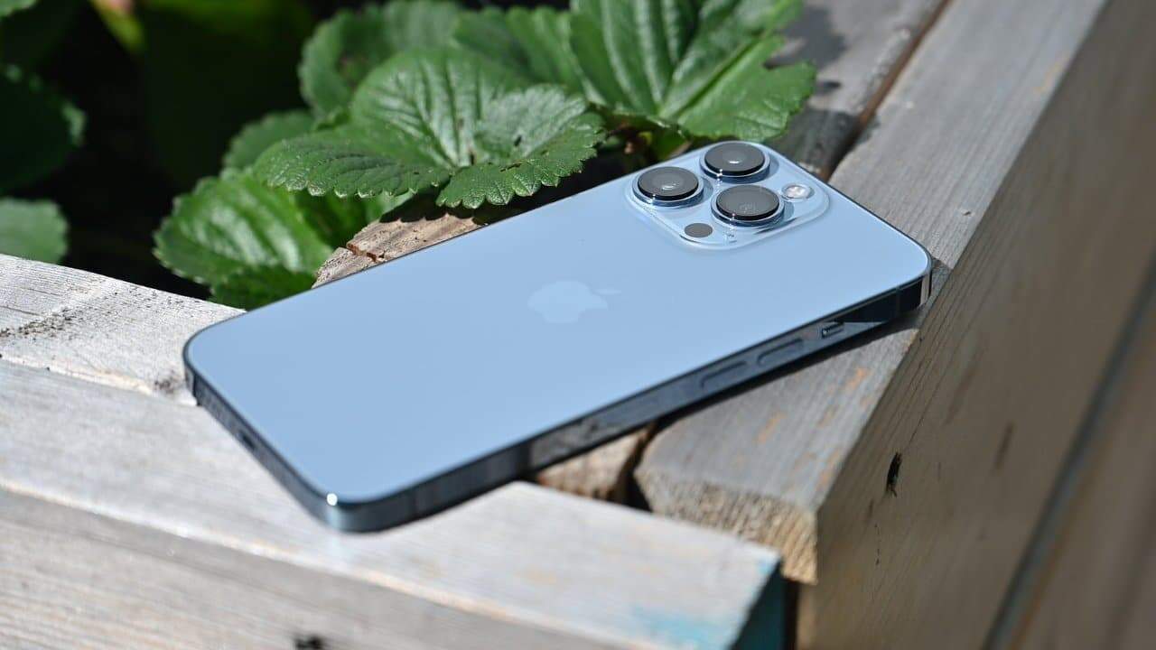 A silver smartphone with three camera lenses placed on a wooden surface near green leaves.