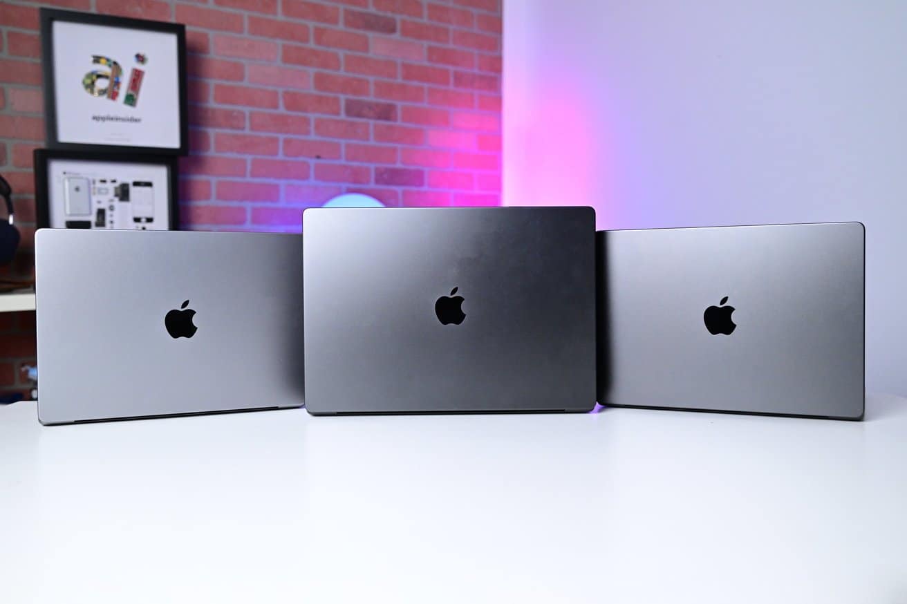 Three MacBook Pros with Apple logos on a white table, against a red brick wall backdrop. Pink and blue lighting illuminate the scene.