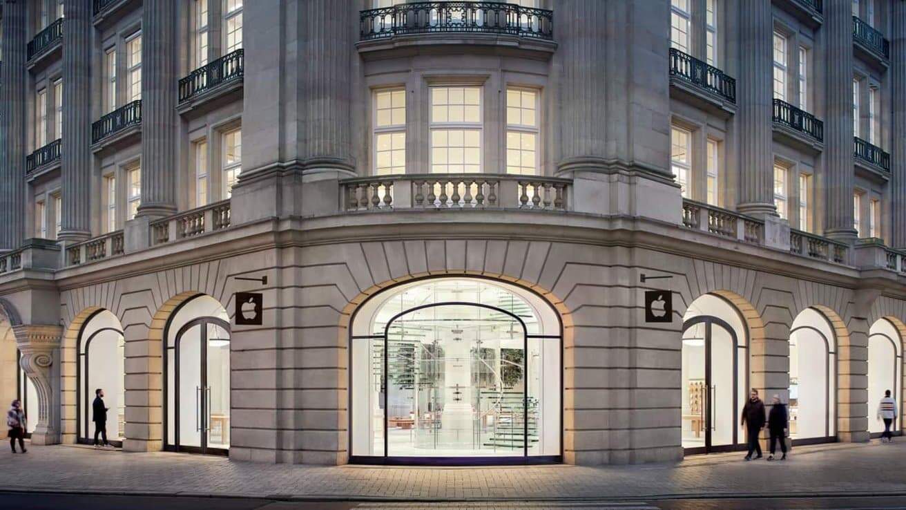 Elegant building with tall windows, stone columns, and arched doorways featuring modern glass entrance. A logo sign hangs outside. People walk near the store entrance.