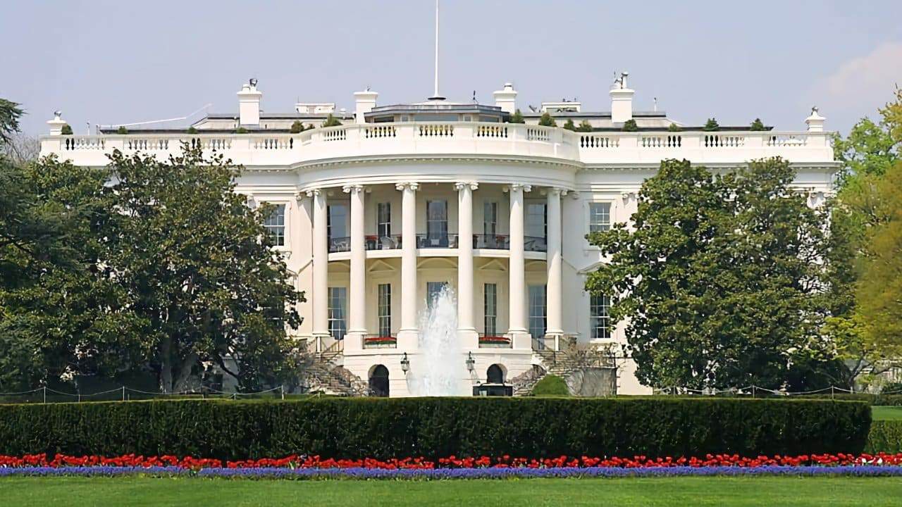 White building with columns, surrounded by trees, a fountain in front, and manicured gardens with red and purple flowers.