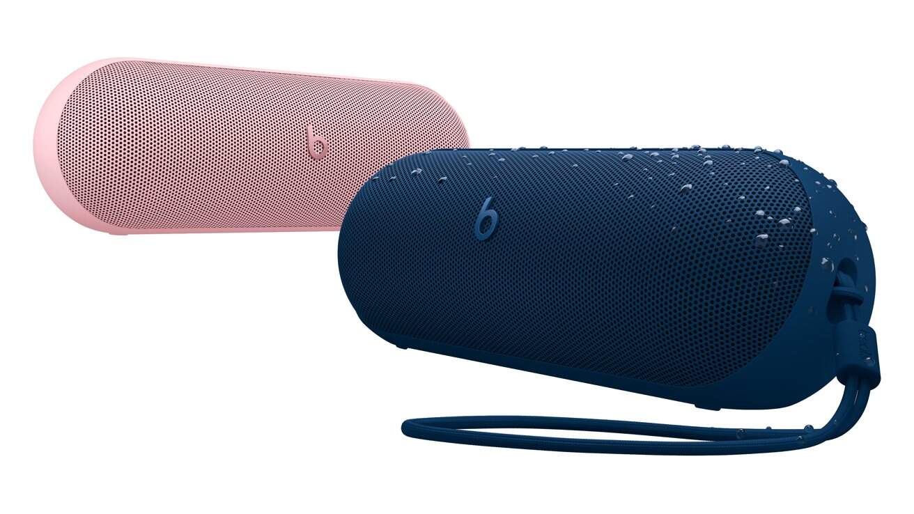 Two portable wireless speakers, one pink and one blue. The blue speaker has water droplets on it and a carrying strap. Both feature a perforated mesh design with a 'b' logo.