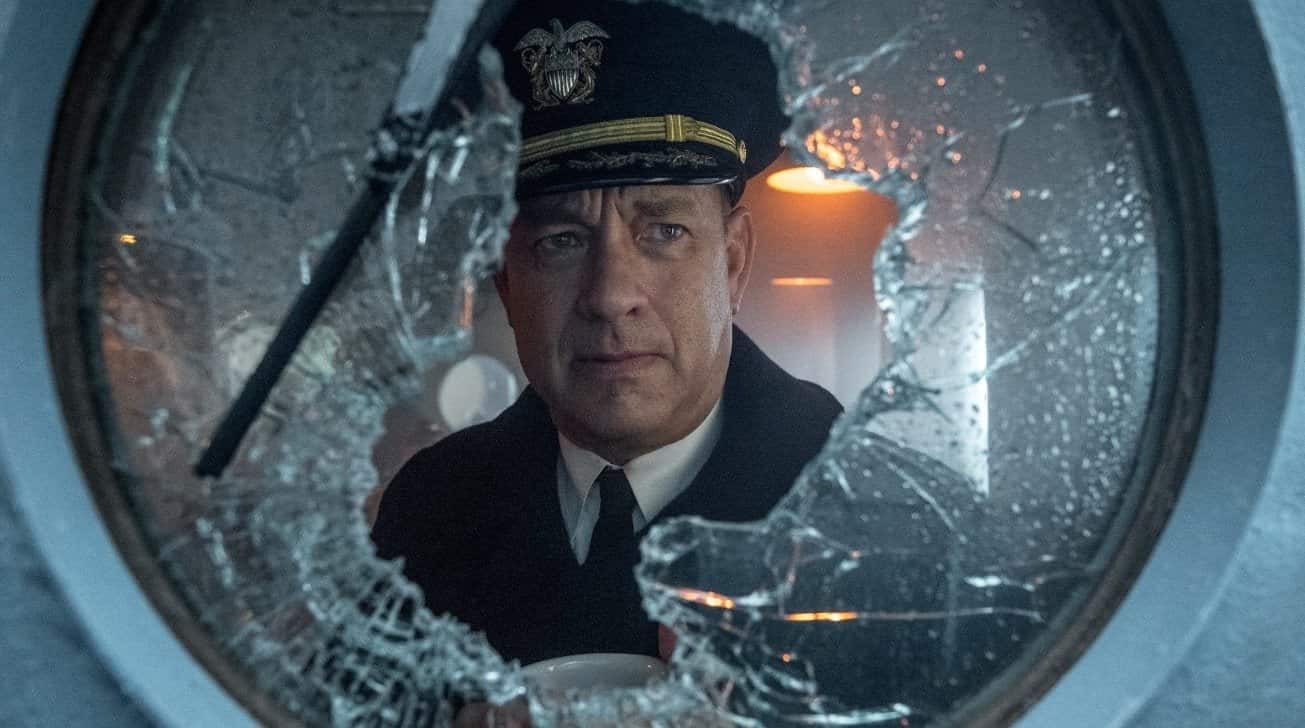 Man in a navy uniform stands behind a shattered glass window, looking serious and holding a cup, with a dimly lit background.
