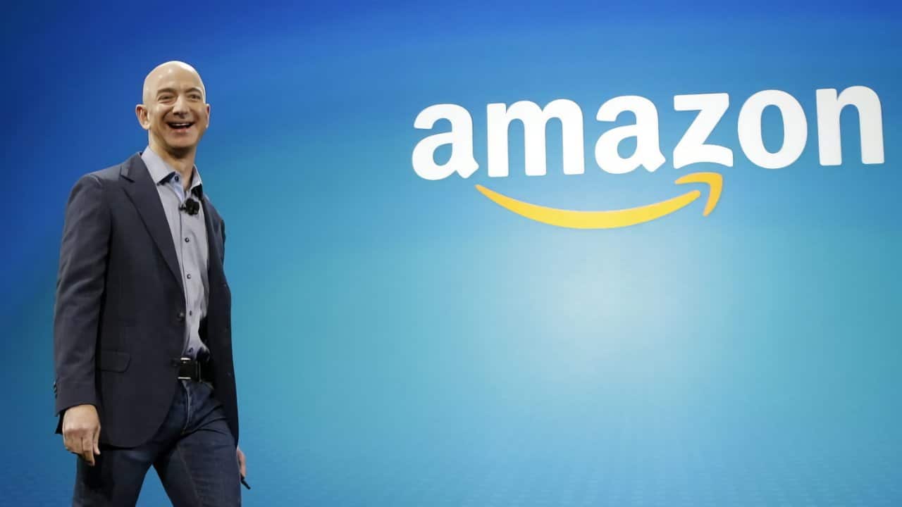 Smiling man in a suit stands in front of a blue background with the Amazon logo, featuring the word 'amazon' and a yellow arrow.