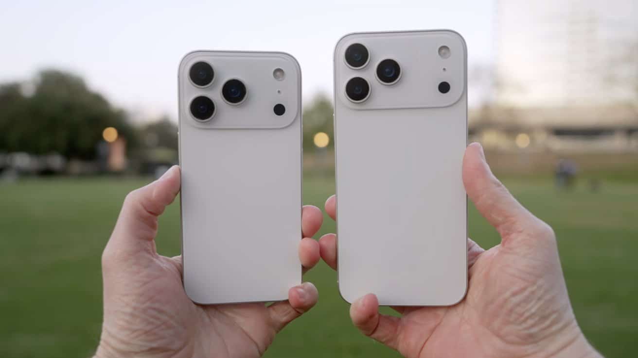 Two hands holding white smartphones with triple cameras against a grassy park background.