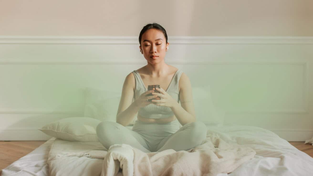 A person sits cross-legged on a bed, eyes closed, holding a small jar, in a calm, softly lit room.