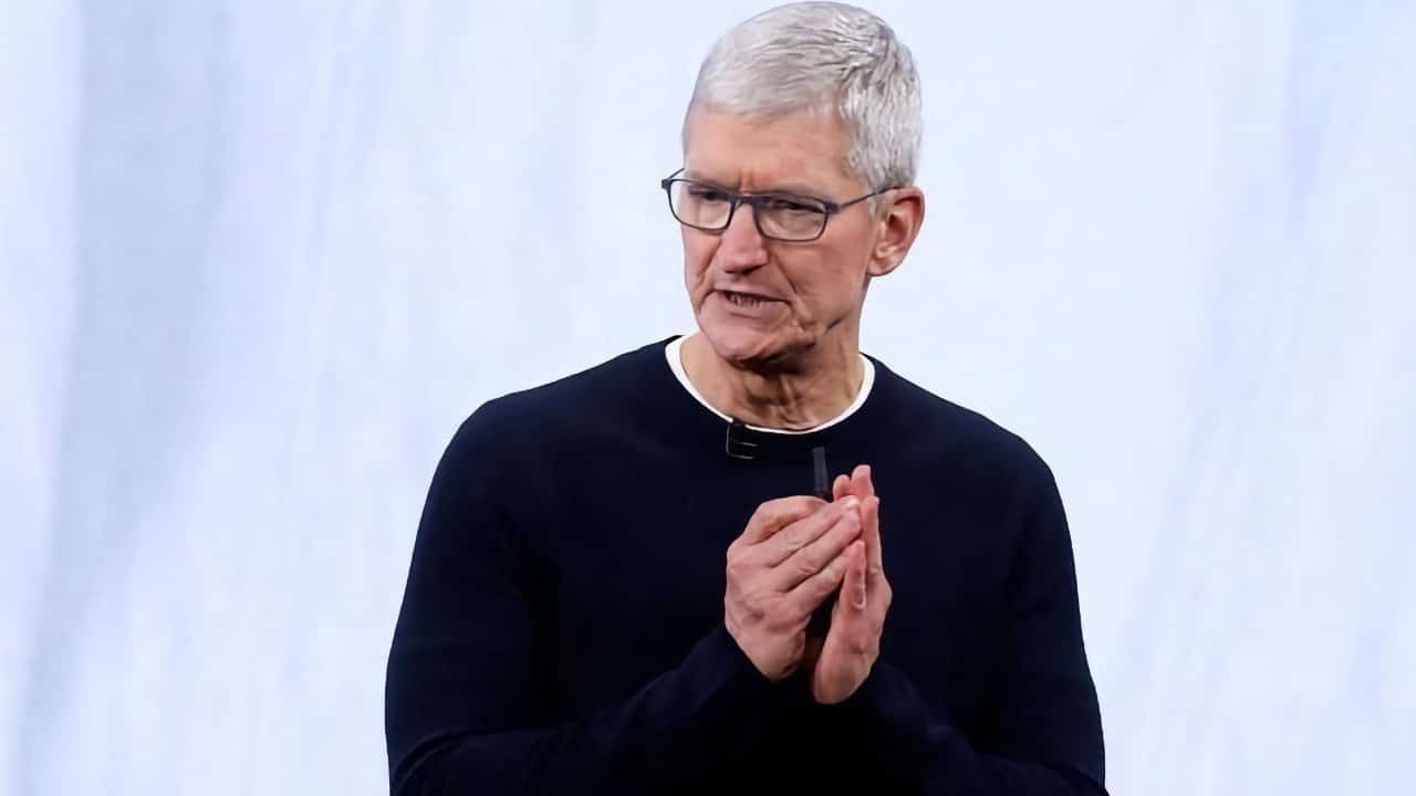 Man with gray hair and glasses, wearing a black shirt, clasping hands, speaking against a plain light background.
