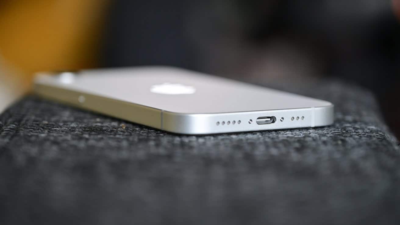 Close-up of a silver iPhone 16e placed on a textured surface, focusing on the charging port and speaker holes on the bottom edge.