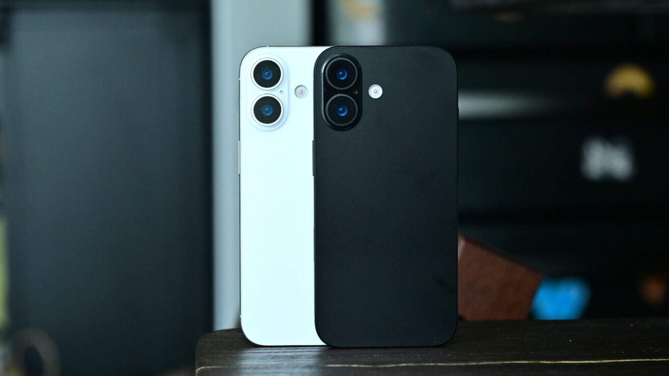 Two smartphones, one white and one black, with dual camera lenses, standing upright on a wooden surface against a dark blurred background.