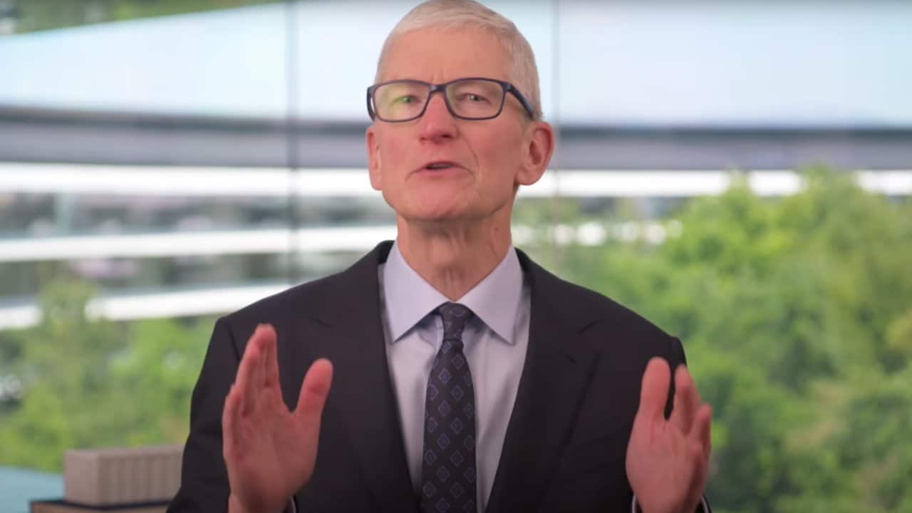 Man in a suit gestures while speaking, set against a blurred backdrop of greenery and a modern building with large windows.