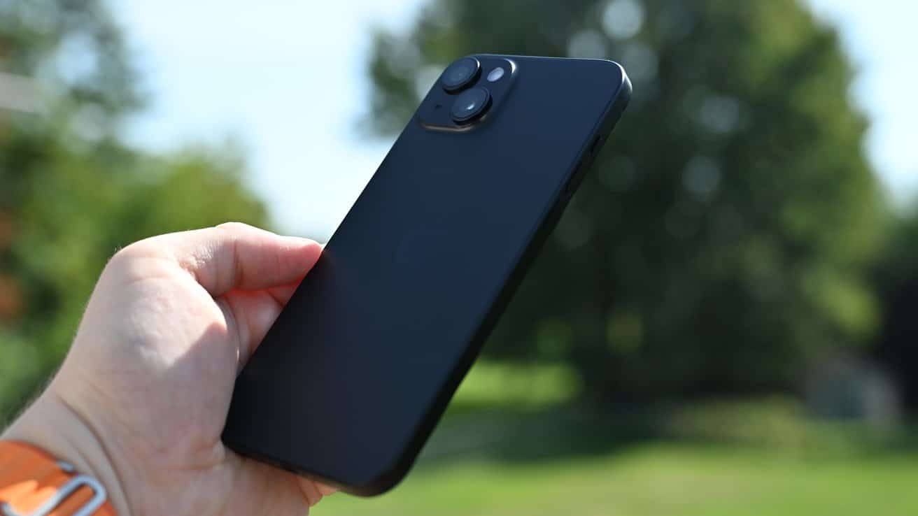 Hand holding a black iPhone 15 with dual cameras outdoors, green blurred background.