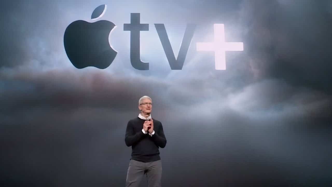 Person standing on stage with dark clothes, hands clasped, in front of a cloudy background displaying a large Apple TV+ logo.
