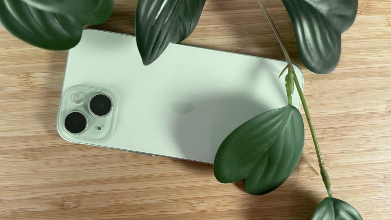 Smartphone with dual camera lenses on a light wooden surface, surrounded by large, dark green leaves.
