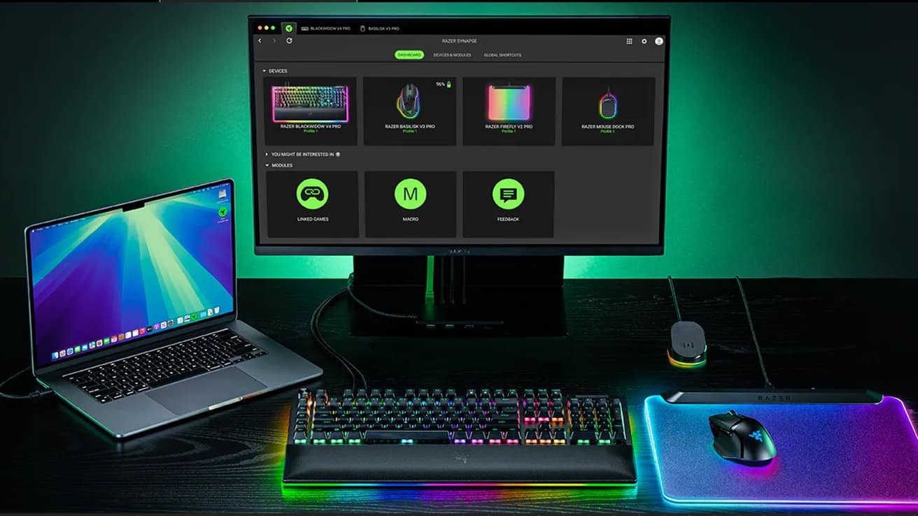 Modern gaming setup featuring a colorful backlit keyboard, mouse on a glowing pad, large monitor displaying software interface, and a laptop with a vibrant screen.