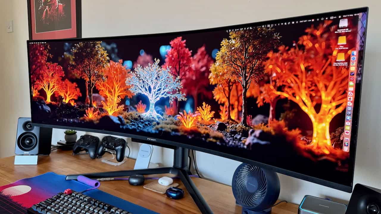 Ultrawide monitor displaying a vibrant digital forest scene with glowing orange and white trees, on a desk with speakers, gaming controllers, keyboard, and small fan.