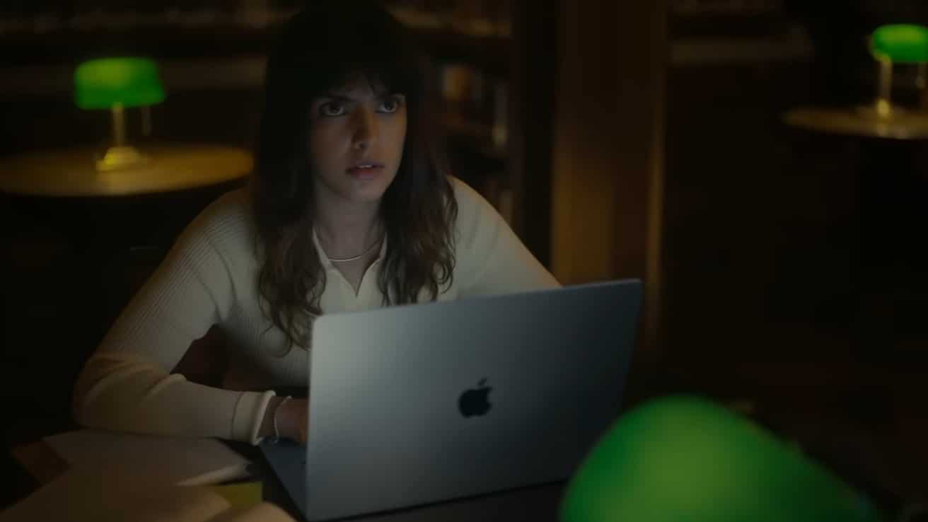 Woman with long hair working on a laptop in a dimly lit room, illuminated by green desk lamps, appears thoughtful.