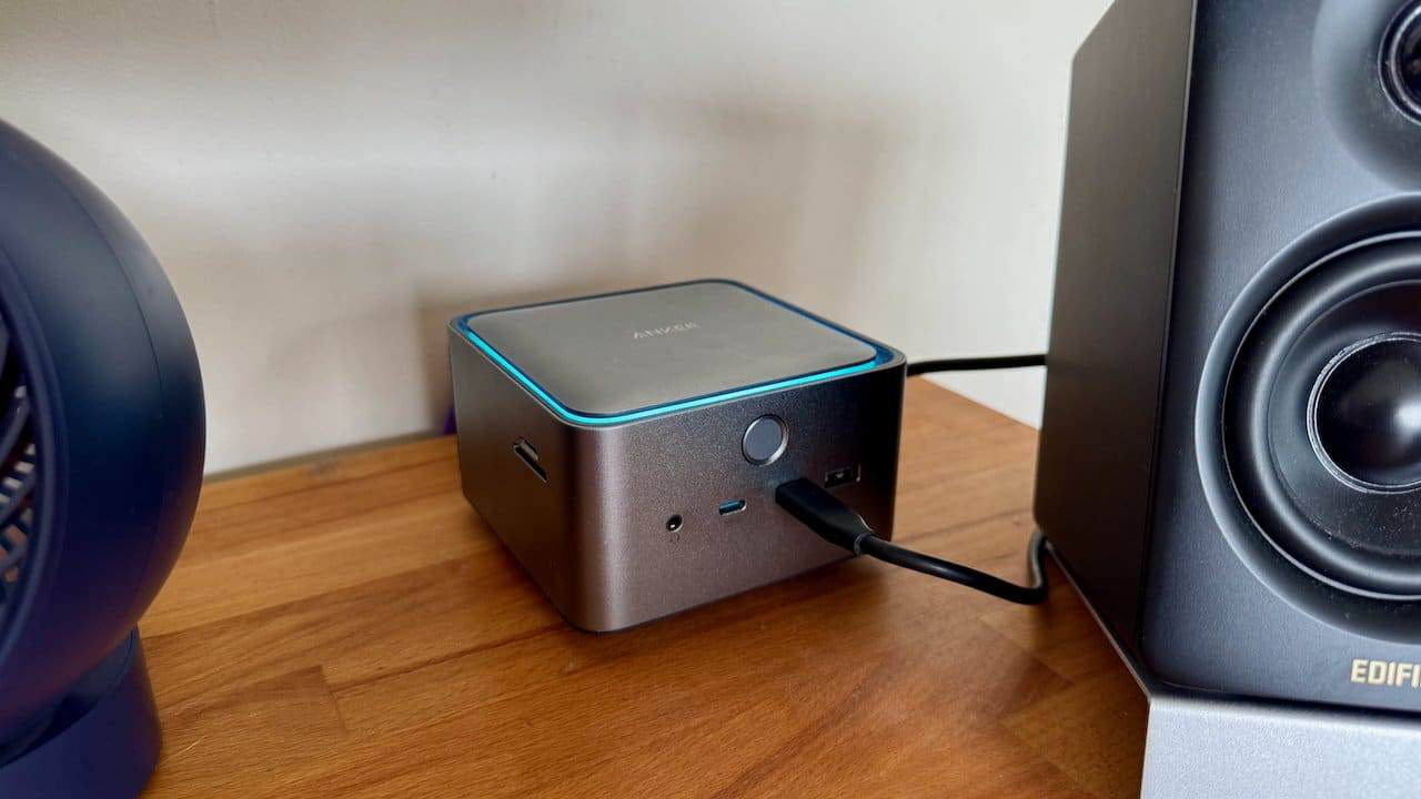 Anker Prime Thunderbolt 5 dock with blue accent lighting on a wooden desk, connected by a cable to nearby black speakers.
