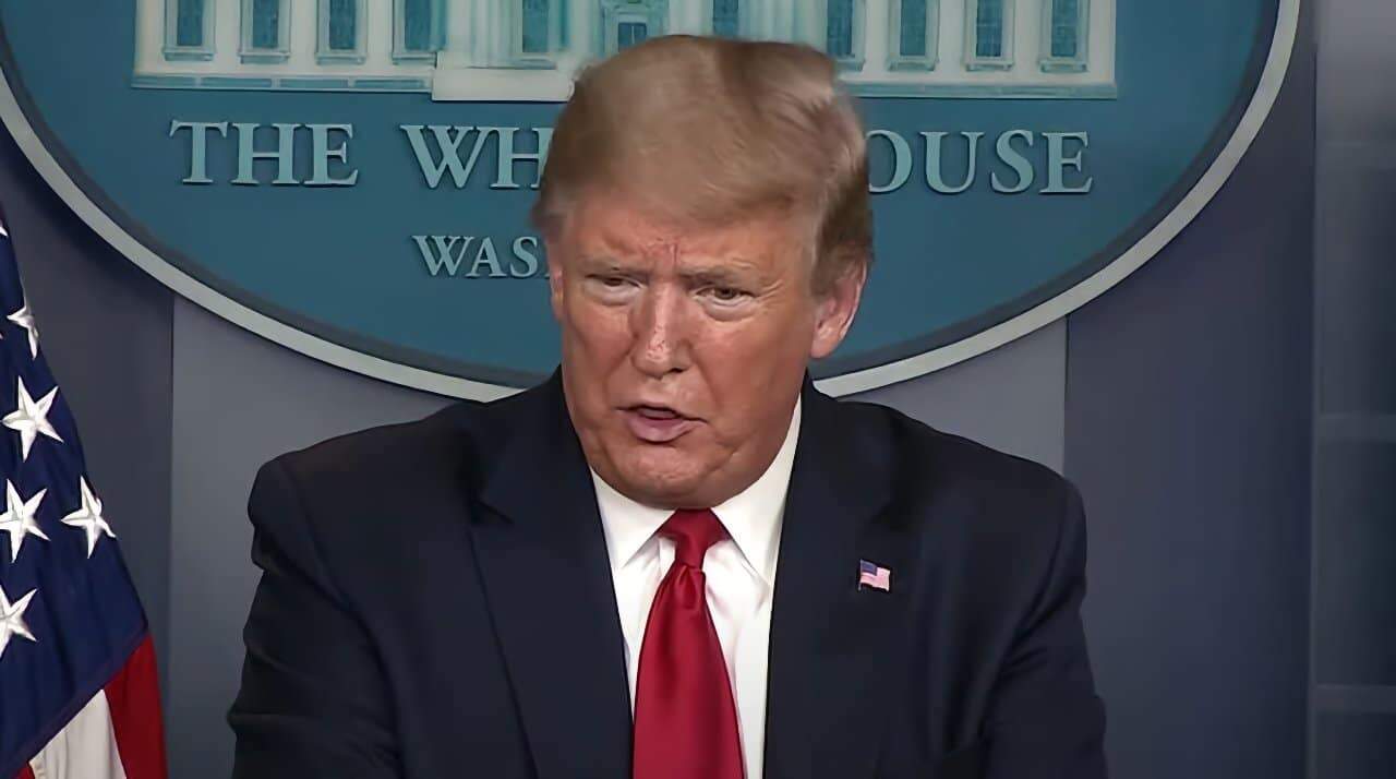 A man in a suit speaks at a podium in front of a blue background with 'The White House' written on it, American flag partially visible.