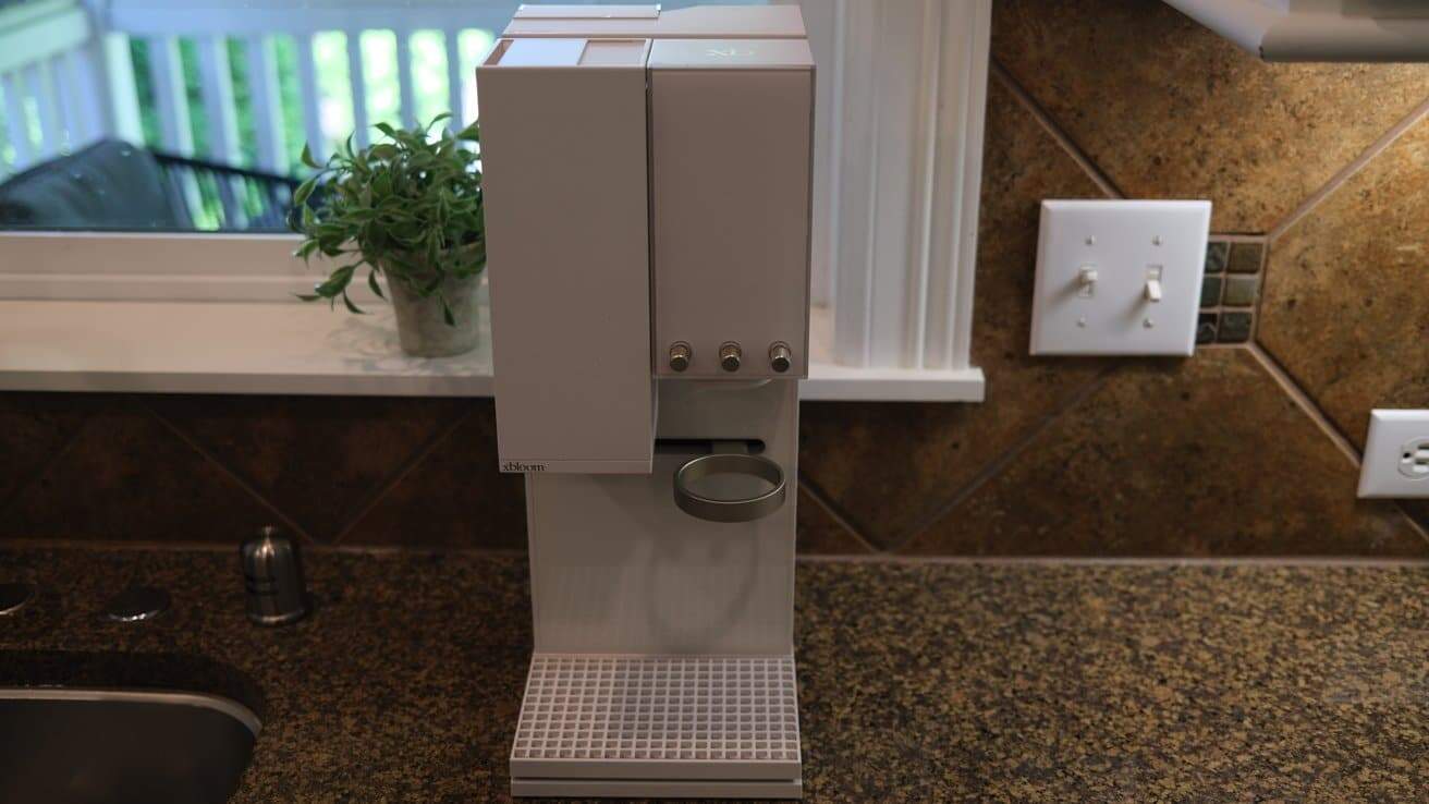 White espresso machine on a granite countertop, surrounded by a sink, potted plant, and light switches, with a window in the background.