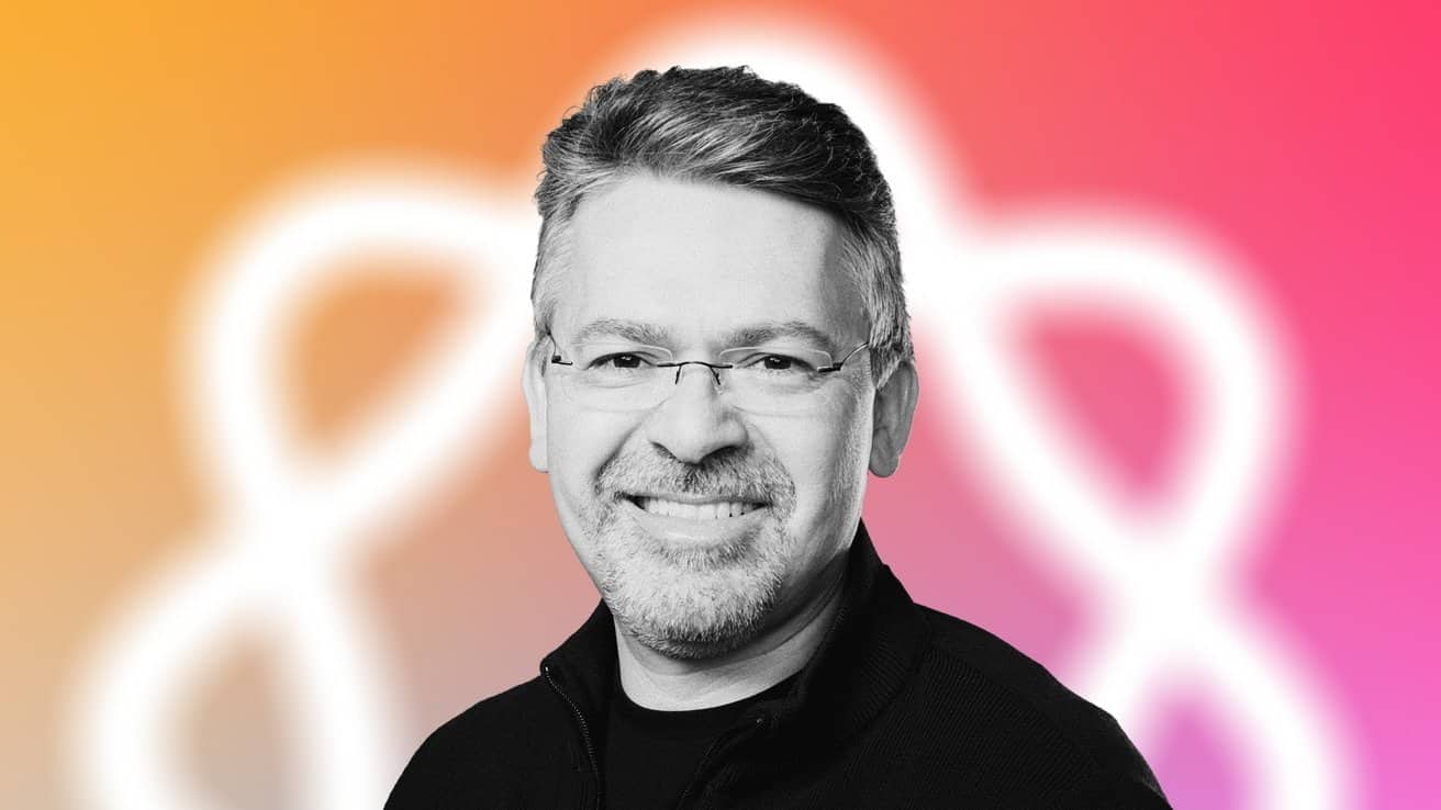 Smiling man with glasses, beard, and short hair against a vibrant orange to pink gradient background featuring a blurred white, abstract shape.