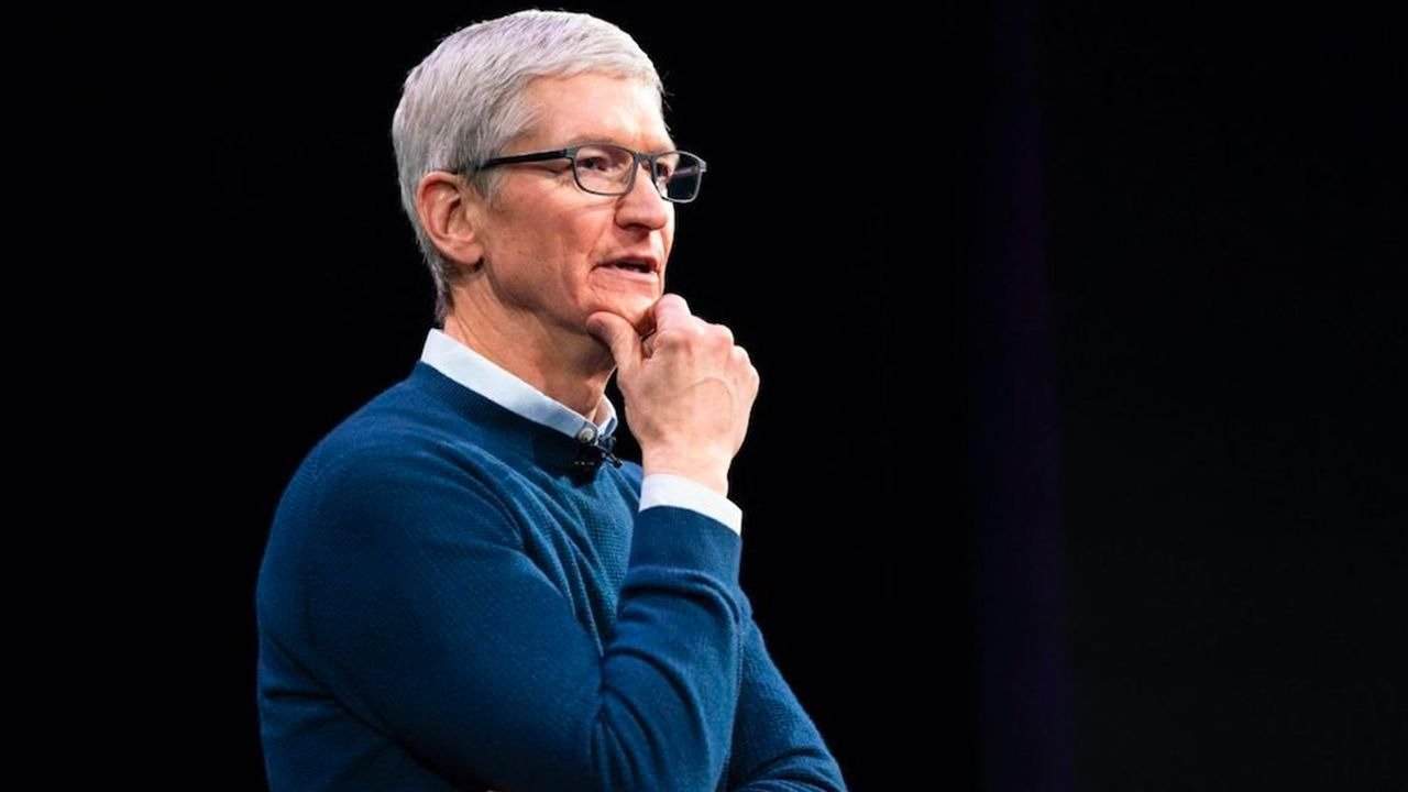 Tim Cook with short gray hair, wearing glasses and a blue sweater, poses thoughtfully against a dark background.