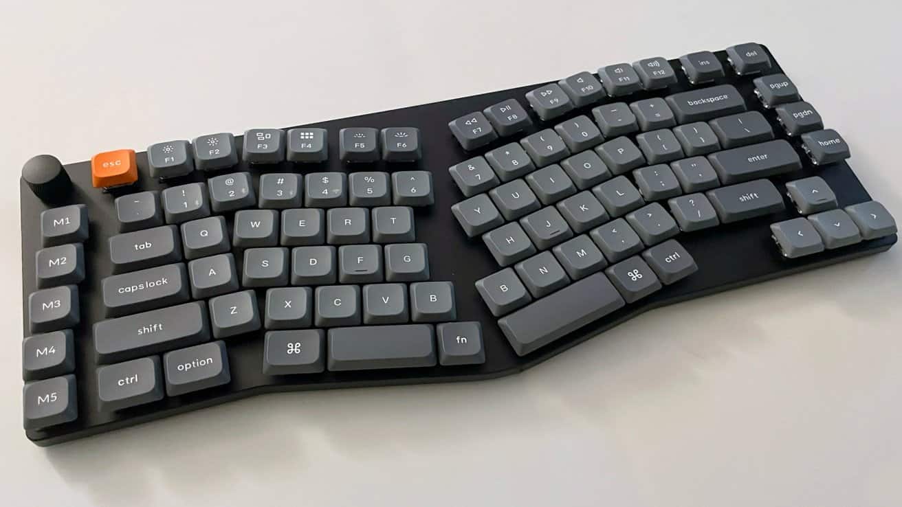 Ergonomic mechanical keyboard with split design, dark gray keys, orange escape key, and extra function keys on the left side.