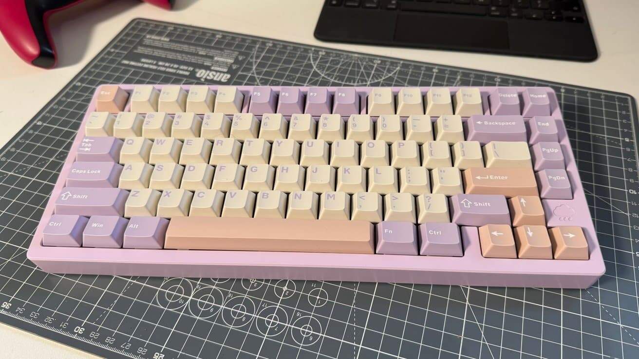 Purple and beige keyboard with mixed keycap colors on a cutting mat surface, accompanied by a red and black game controller and a laptop in the background.