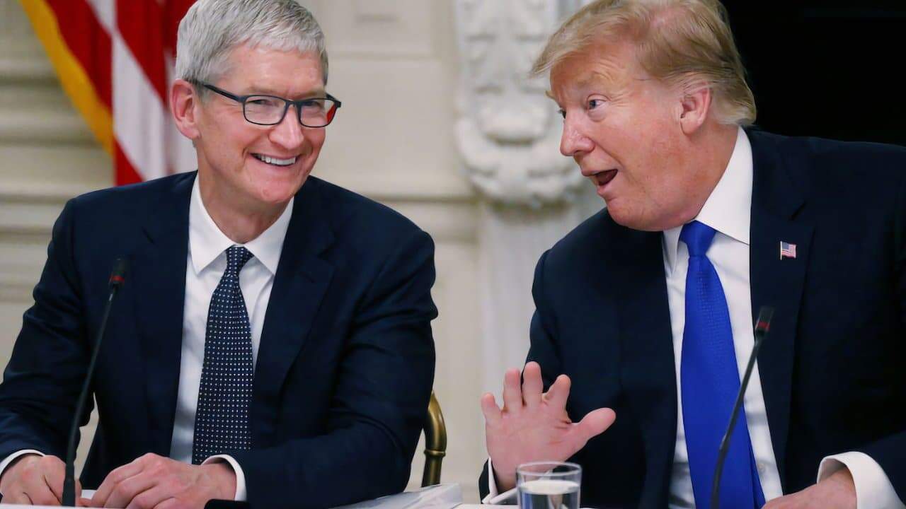 Two men in suits sit at a table, engaged in conversation, with one smiling and the other gesturing animatedly. An American flag is partially visible in the background.
