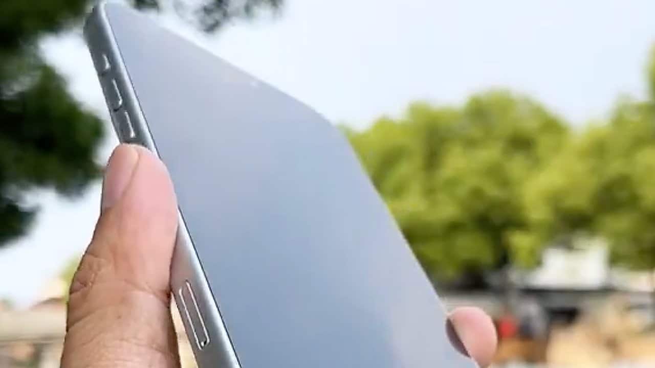 A hand holding a slim silver smartphone outdoors, with green trees blurred in the background.