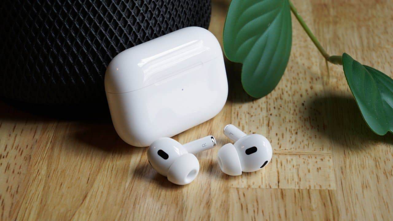 White wireless earbuds and matching case on a wooden surface, alongside a speaker with a textured black mesh and a green leafy plant.