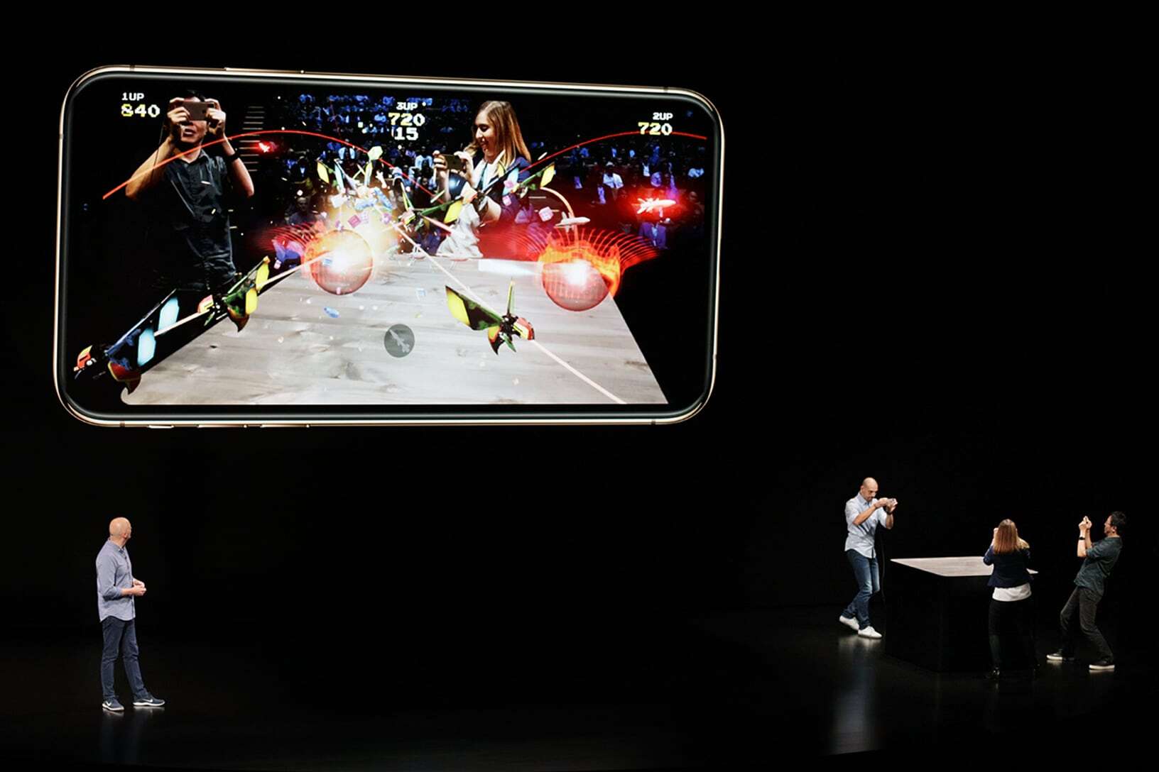 Three people on stage play Galaga AR on iPhones while a fourth looks on.