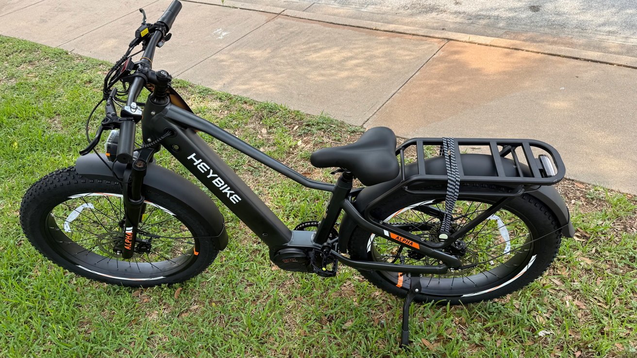 Black electric bike with fat tires, rear cargo rack, and visible branding, parked on grass beside a sidewalk.