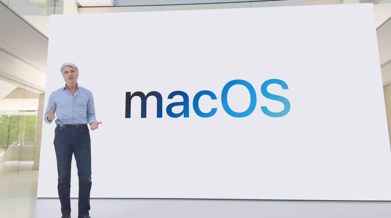 A person in a blue shirt and jeans stands in front of a large screen displaying the word 'macOS'.