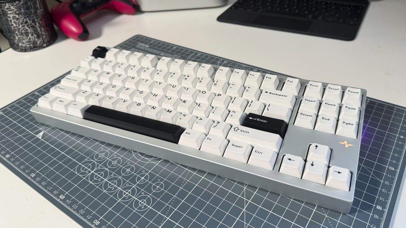 White mechanical keyboard with gray base and black key accents on a grid mat, red game controller and black tablet in the background.