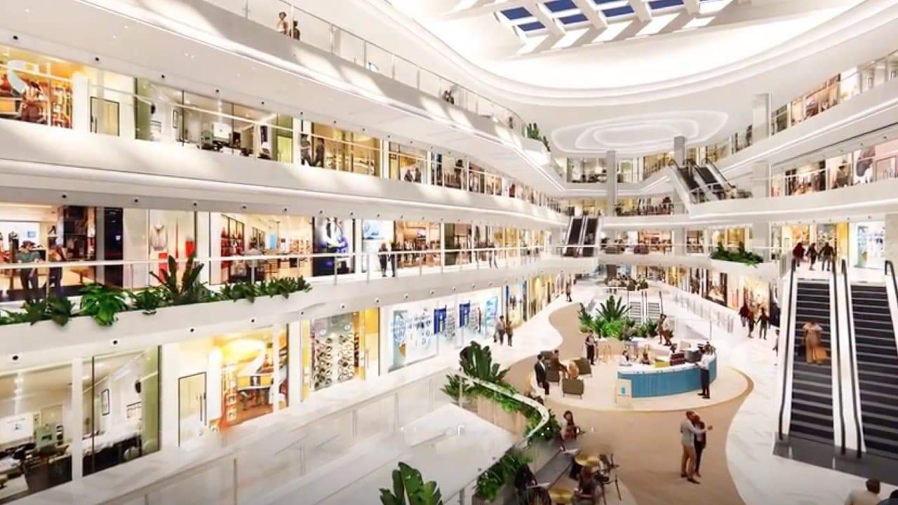 Spacious multi-level shopping mall with escalators, bright lighting, indoor plants, and people exploring various shops and cafes. Skylights illuminate the open, modern design.