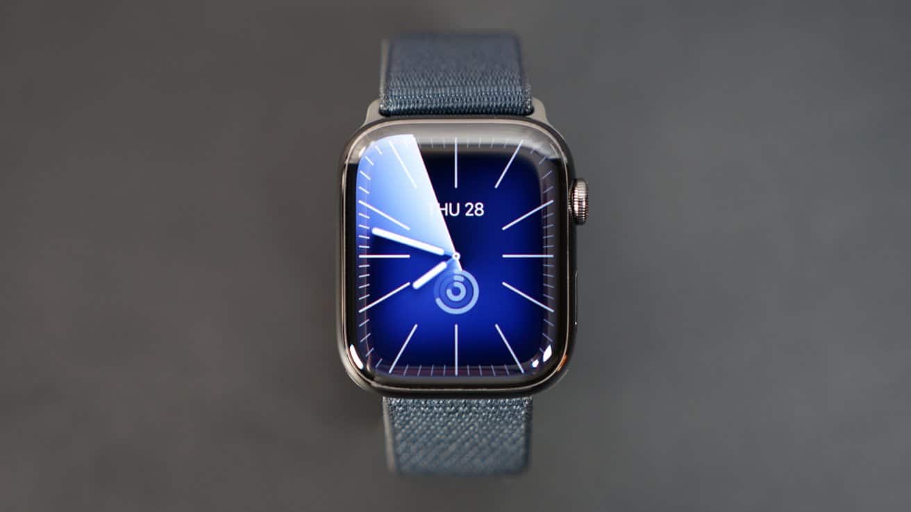 Smartwatch with a blue face displaying time and date, attached to a blue fabric strap against a dark background.