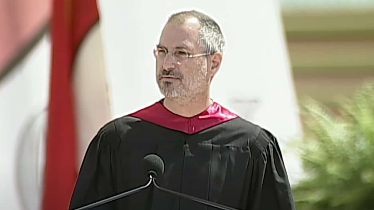 Person in black and red academic gown speaking at an outdoor event, with foliage and a blurred background.