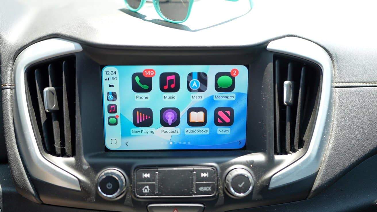 Car infotainment screen displaying various app icons, such as Phone, Music, Maps, and Messages, with notifications. Sunglasses rest on top of the dashboard.