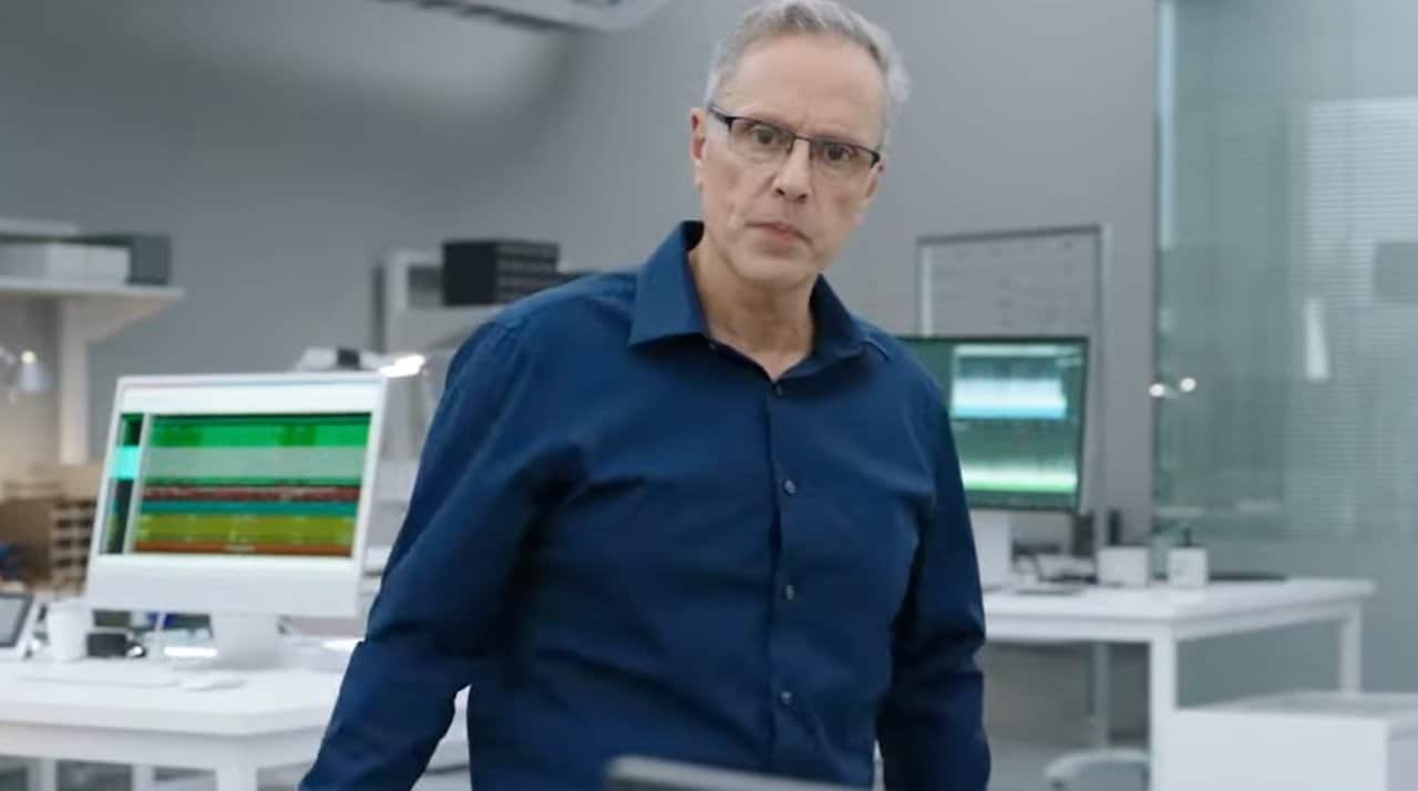 Older person in glasses wearing a blue shirt, standing in a modern office with computers in the background.