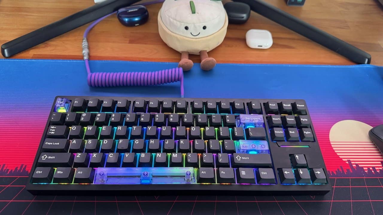RGB mechanical keyboard on neon blue and pink mat, coiled purple cable, stuffed toy, and tech gadgets on wooden desk.