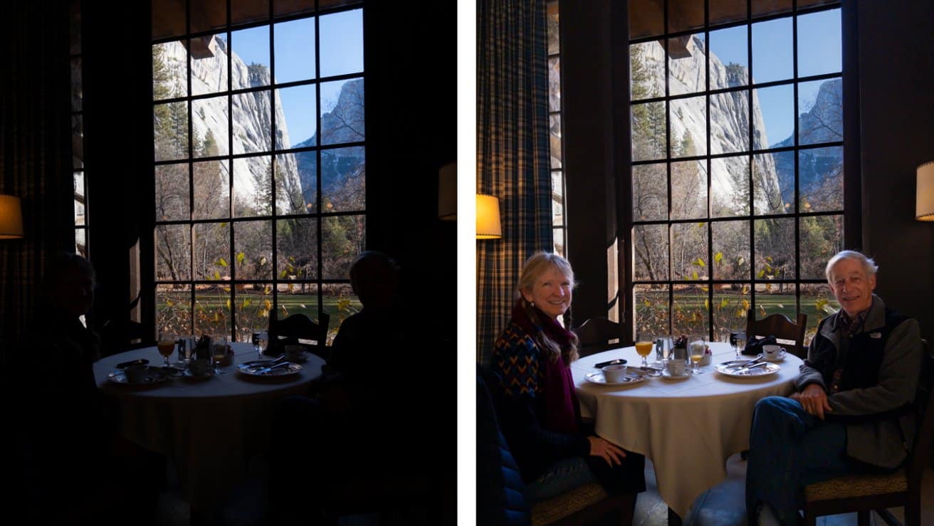 Two people sitting at a round table with breakfast, in front of a large window showing a scenic mountain view. The room is softly lit.